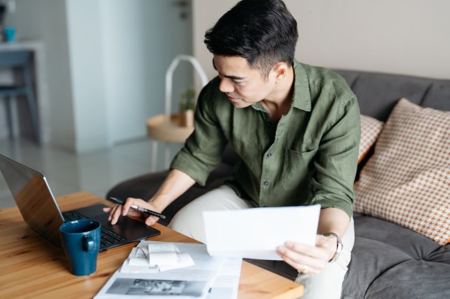 Asian man using laptop and holding receipts while managing finances at home